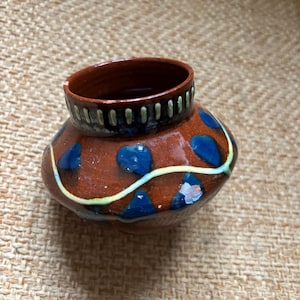 Puede incluir: Una pequeña olla de cerámica hecha a mano con un cuerpo redondeado y una parte superior ancha y abierta. La olla es principalmente marrón con formas azules irregulares y una fina línea amarilla. El borde es marrón oscuro con un patrón negro y verde.