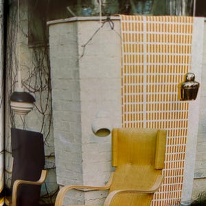 May include: An interior scene featuring a light wood chair with a curved design, a dark chair, and hanging gold-colored decorative objects. A patterned wall hanging with vertical lines in yellow and white is also visible.