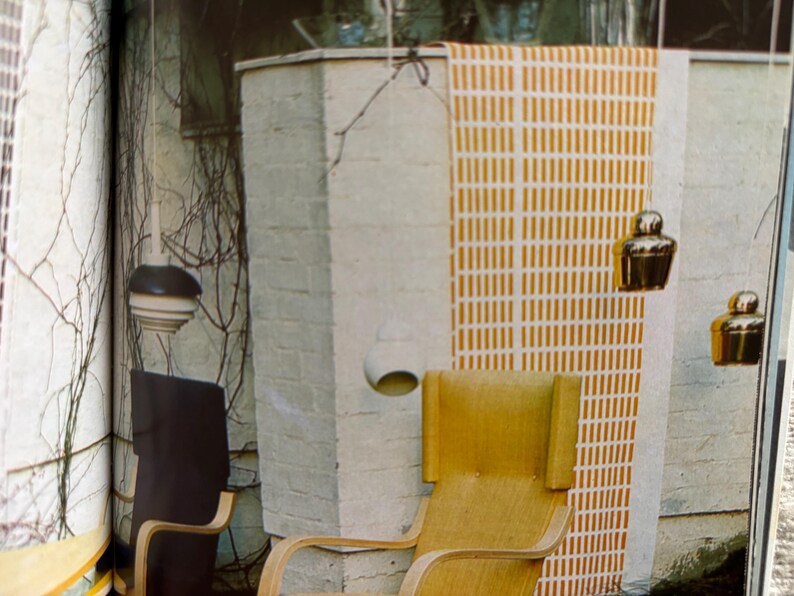 May include: An interior scene featuring a light wood chair with a curved design, a dark chair, and hanging gold-colored decorative objects. A patterned wall hanging with vertical lines in yellow and white is also visible.