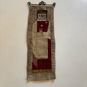 May include: A hand-woven wall hanging featuring a stylized figure in a red and white robe, holding a staff. The figure is embroidered on a beige burlap background.