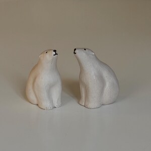 Op de afbeelding: Twee kleine, witte ijsbeerbeeldjes. De beren zitten rechtop, met hun hoofden omhoog gekanteld. De beeldjes hebben een gladde, matte afwerking en staan op een lichtgekleurd oppervlak.