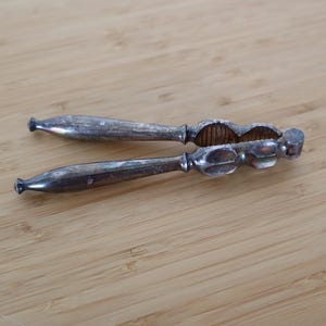 Könnte beinhalten: Ein silberfarbener Nussknacker aus Metall mit zwei länglichen Griffen, die zu den Enden hin schmaler werden, und einem zentralen Klappmechanismus mit einer texturierten, geriffelten Innenseite zum Greifen und Knacken von Schalen.