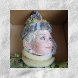 May include: A porcelain doll with blonde hair wearing a camouflage hat and a white foam neck brace. The doll has green eyes and is looking up.