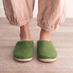 May include: A pair of green felt slippers with a light tan sole. The slippers are a solid green colour and appear soft and comfortable. The image shows the slippers from a front-facing perspective.