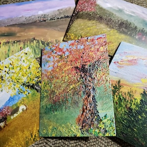 May include: Five different paintings on cards, each depicting a different landscape scene. The first card shows a sunset over a field with a small town in the distance. The second card shows a field of flowers with a tree in the background. The third card shows a close-up of a tree with red and orange leaves. The fourth card shows a field of grass with a mountain in the distance. The fifth card shows a field of grass with a sunset in the background.