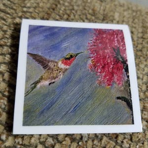 May include: A painting of a hummingbird in flight, with a red head and green body, hovering near a cluster of pink flowers. The background is a blue and green wash of color.