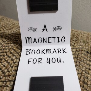 May include: A white card with black text that reads "A Magnetic Bookmark For You." Two black rectangular magnets are attached to the card.