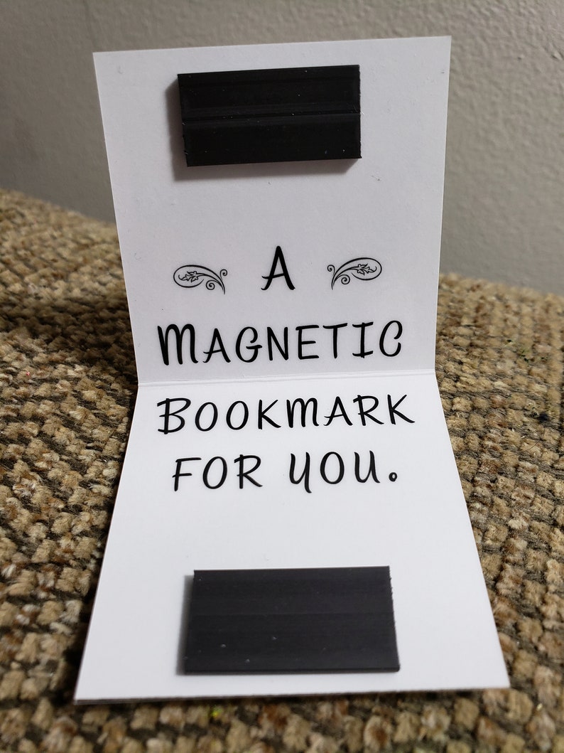 May include: A white card with black text that reads "A Magnetic Bookmark For You." Two black rectangular magnets are attached to the card.