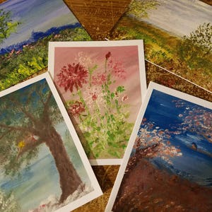 May include: Five painted cards with different nature scenes. The cards are on a wooden surface. The scenes include a blue sky with mountains, a field of pink and white flowers, a tree with a bird's nest, a blue sky with a tree and a bird, and a field with a tree and a blue sky.