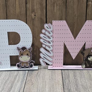May include: Wooden letter cutouts, one white with the name "Paisley" in pink script and a horse graphic, and one pink with the name "Millie" in white script and a Highland cow graphic. The letters are dotted and stand on white bases.