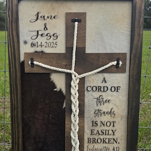 May include: A framed wooden sign with a cowhide background and a wooden cross overlay. The sign includes the names "Jane & Jess" and the date "6-14-2025". Text on the sign reads "A cord of three strands is not easily broken. Ecclesiastes 4:12."