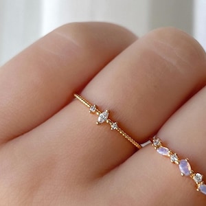 May include: Two delicate gold rings, each set with small gemstones, are shown on a hand. One ring has a line of small, clear stones, the other features a series of iridescent, oval-shaped stones and clear accents. The background is a soft, neutral colour.