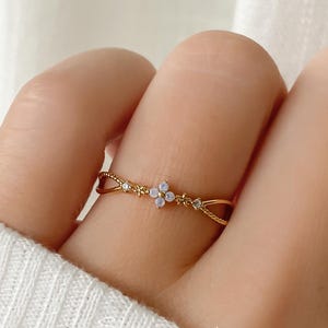 以下が含まれることがあります： 繊細なデザインのゴールドカラーのリング。リングの中央には、小さな淡色の石が十字型に集まっており、バンドに沿って小さな透明な石と織り込まれたディテールが施されています。リングは指に着用されています。