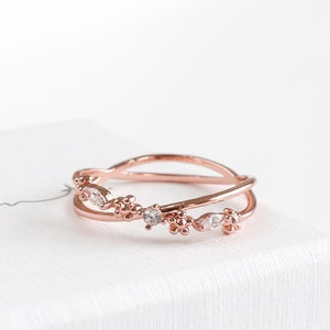 Peut inclure: Une bague en or rose avec un design délicat et entrelacé orné de petites pierres précieuses claires. La bague est présentée sur une boîte blanche avec le texte "Statement Grey" écrit en cursive.