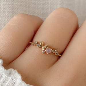 Könnte beinhalten: Ein zarter Goldring mit einem rosafarbenen Stein und kleinen klaren Steinen, die in einem floralen Design eingefasst sind.