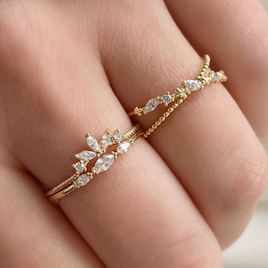 May include: Two gold rings, one with a leaf-like arrangement of clear gemstones and the other a simple band with smaller stones. The rings are stacked on a finger. The rings are made of gold and feature clear gemstones.