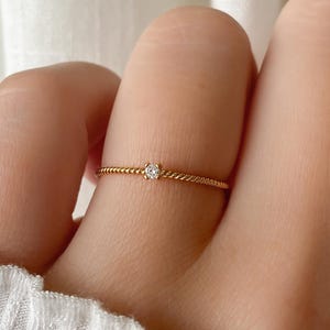 May include: A delicate gold ring with a twisted band and a small, clear gemstone at the center. The ring is worn on a finger, showcasing its fine details and elegant design. The ring's band has a textured appearance.