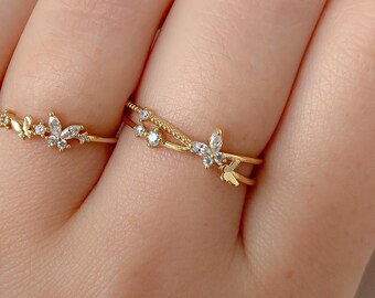 May include: Two gold-colored rings with delicate butterfly and leaf designs. One ring features a curved band with small butterflies and clear gemstones. The other ring has a double band with a butterfly and gemstones.