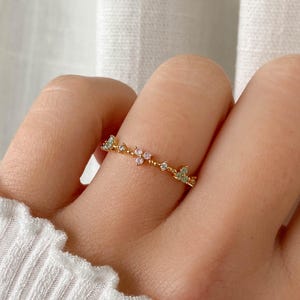 May include: A gold ring featuring a floral design with pink and green gemstones. The ring is worn on a finger and has a delicate, elegant appearance. The ring's design is suitable for everyday wear and is a subtle piece of jewellery.