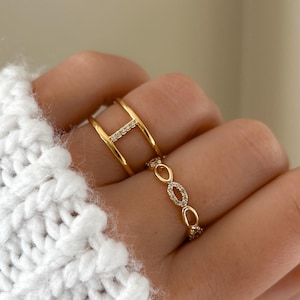 May include: Two gold rings set with clear cubic zirconia stones. One ring is a simple band with a row of stones, and the other ring is a thicker band with a geometric design.