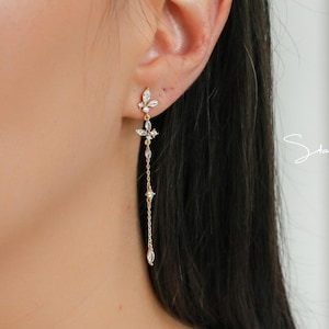 May include: Gold-toned drop earrings with a floral design and dangling teardrop-shaped accents. The earrings are worn on an ear, showcasing their delicate chain and sparkling details. The earrings are a statement piece.