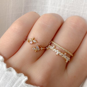 May include: A set of three gold rings with delicate leaf designs and clear and pink gemstones. The rings are stacked on a finger.
