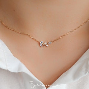 May include: A delicate gold necklace with a small leaf pendant featuring blue and white gemstones. The necklace is on a model wearing a white shirt.