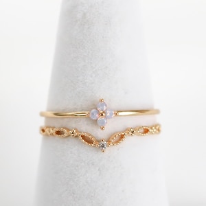 Könnte beinhalten: Zwei goldene Ringe. Der obere Ring hat eine kleine, vierblättrige Blume mit hellblauen Steinen. Der untere Ring hat ein filigranes, offenes Design mit kleinen, klaren Steinen. Beide Ringe werden auf einem weißen, kegelförmigen Ständer präsentiert.