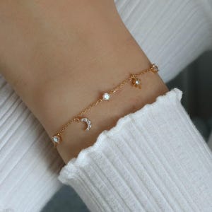 May include: Gold-toned bracelet with dangling charms, including a crescent moon, stars, and round gems. The bracelet is worn on a wrist, against a white ribbed long-sleeve shirt. The word "Statement" and the word "GREY" are written in the image.