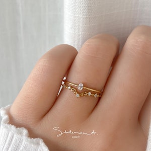 May include: Two gold rings with small clear stones. The top ring has a larger oval-shaped stone. The rings are on a finger.