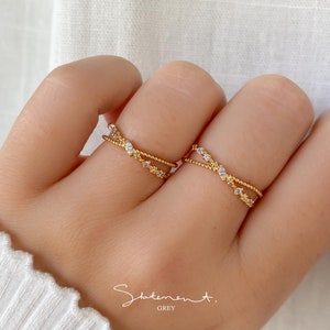 Könnte beinhalten: Zwei goldene Ringe mit kleinen, klaren Steinen, die in einem Kreuzmuster gesetzt sind. Die Ringe werden an zwei Fingern einer Hand getragen.