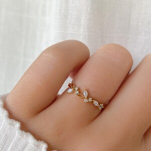 Puede incluir: Un anillo dorado con un diseño de hojas y piedras preciosas. El anillo presenta pequeñas piedras preciosas transparentes y se lleva en un dedo. La palabra "Statement" y la palabra "GREY" están impresas en la parte inferior de la imagen.