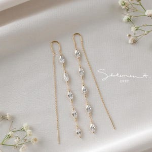 May include: A pair of gold-toned threader earrings with multiple marquise-cut clear crystals. The earrings are displayed on a white surface with floral accents and the text "Statement. GREY".