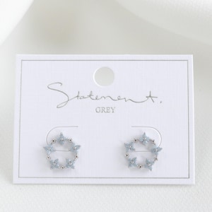 May include: A pair of silver stud earrings with a circular design featuring light blue gemstones. The earrings are displayed on a white card with the text "Statement. GREY".