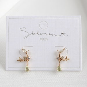 May include: A pair of gold earrings with a delicate floral design and green gemstone accents. The earrings are displayed on a white card with the text "Statement. GREY".