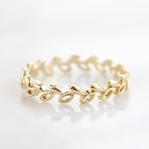 May include: A gold ring with a repeating leaf pattern. The ring's design features small, detailed leaves, creating a nature-inspired look. The ring is displayed on a white surface, emphasizing its intricate details. The ring is a delicate piece of jewellery.