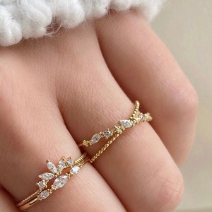 May include: Two gold rings with small clear stones. The top ring has a delicate design of small leaves with stones. The bottom ring has a simple band with small stones.