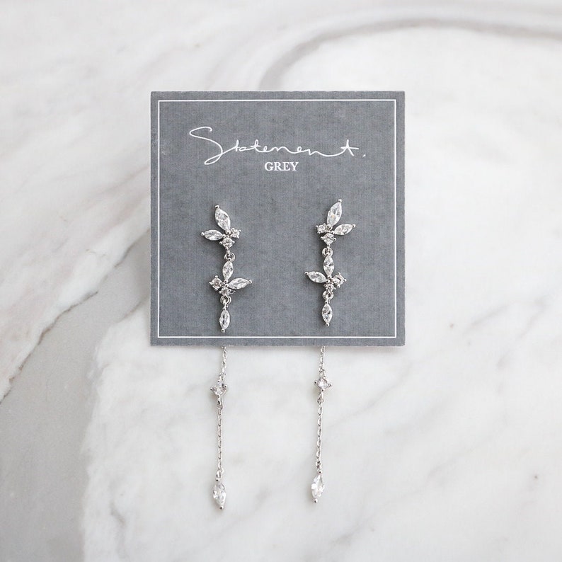 May include: A pair of silver dangle earrings with a floral design and a chain drop. The earrings are on a gray card with the text "Statement. GREY".