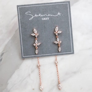 May include: Rose gold dangle earrings with a floral design and clear crystals. The earrings are on a gray card with the text "Statement. GREY".