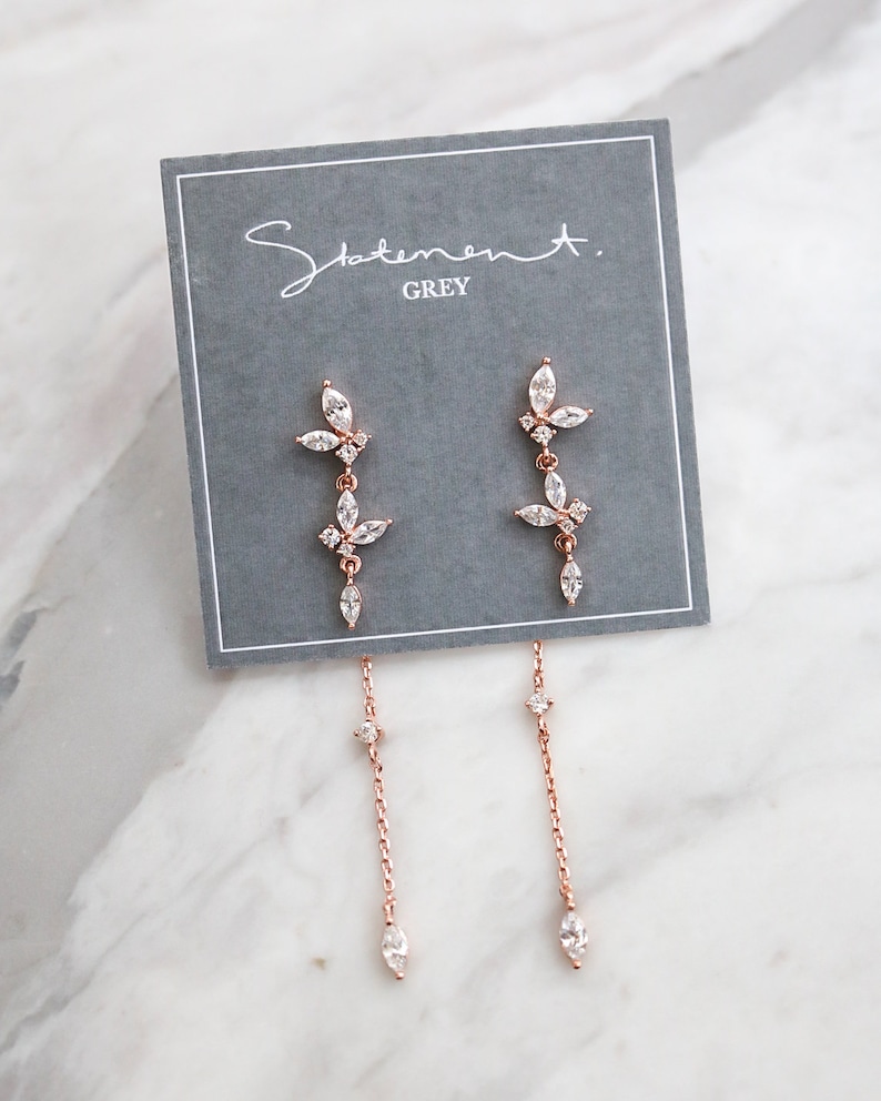 May include: Rose gold dangle earrings with a floral design and clear crystals. The earrings are on a gray card with the text "Statement. GREY".