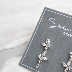 May include: A pair of silver dangle earrings with a floral design featuring clear cubic zirconia stones. The earrings are displayed on a gray card with the text "Statement Grey" printed on it.