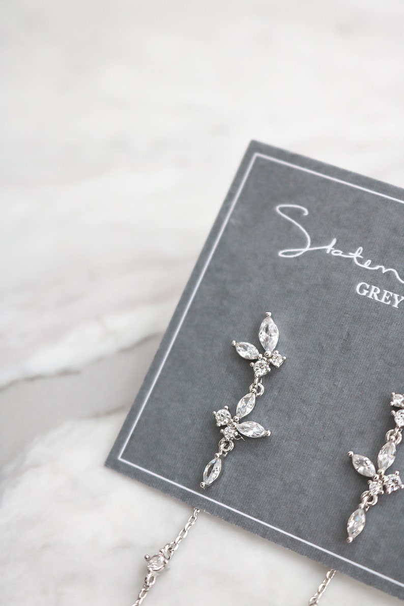 May include: A pair of silver dangle earrings with a floral design featuring clear cubic zirconia stones. The earrings are displayed on a gray card with the text "Statement Grey" printed on it.