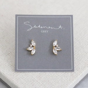 May include: A pair of gold earrings with a cluster of clear cubic zirconia stones in a leaf shape. The earrings are on a grey card with the text "Statement. GREY".