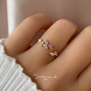 May include: Gold ring with a butterfly design featuring clear and purple gemstones. The ring is open and adjustable.