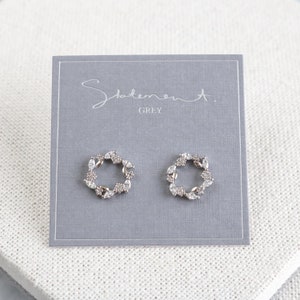 May include: A pair of silver earrings with a circular design featuring small, clear stones. The earrings are displayed on a grey card with the text "Statement. GREY".
