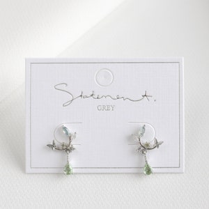 May include: A pair of silver earrings with a delicate floral design and green gemstone accents. The earrings are displayed on a white card with the text "Statement. GREY".