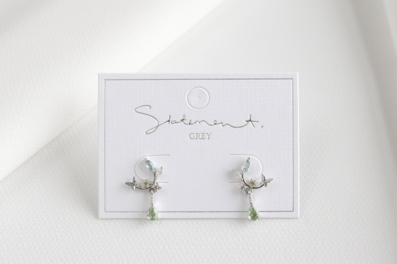 May include: A pair of silver earrings with a delicate floral design and green gemstone accents. The earrings are displayed on a white card with the text "Statement. GREY".