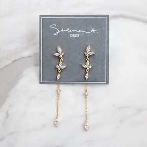 May include: A pair of gold dangle earrings with a floral design and clear crystals. The earrings are on a gray card with the text "Statement. GREY".