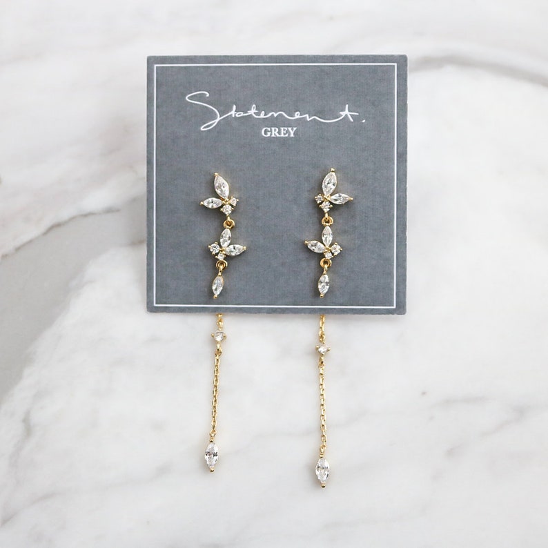 May include: A pair of gold dangle earrings with a floral design and clear crystals. The earrings are on a gray card with the text "Statement. GREY".
