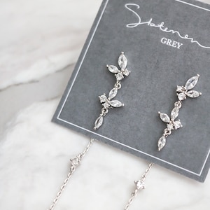 May include: A pair of silver dangle earrings with a delicate chain and a cluster of clear cubic zirconia stones in the shape of leaves. The earrings are displayed on a gray card with the text "Statement Grey".